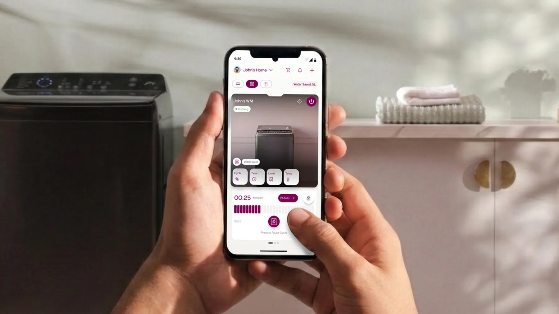 Two hands holding a phone running a smart washing-machine app, with the machine itself beside them in a laundry room